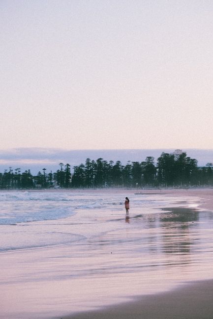 A peaceful scene of a lone person walking along the beach at sunset with gentle waves and distant trees.