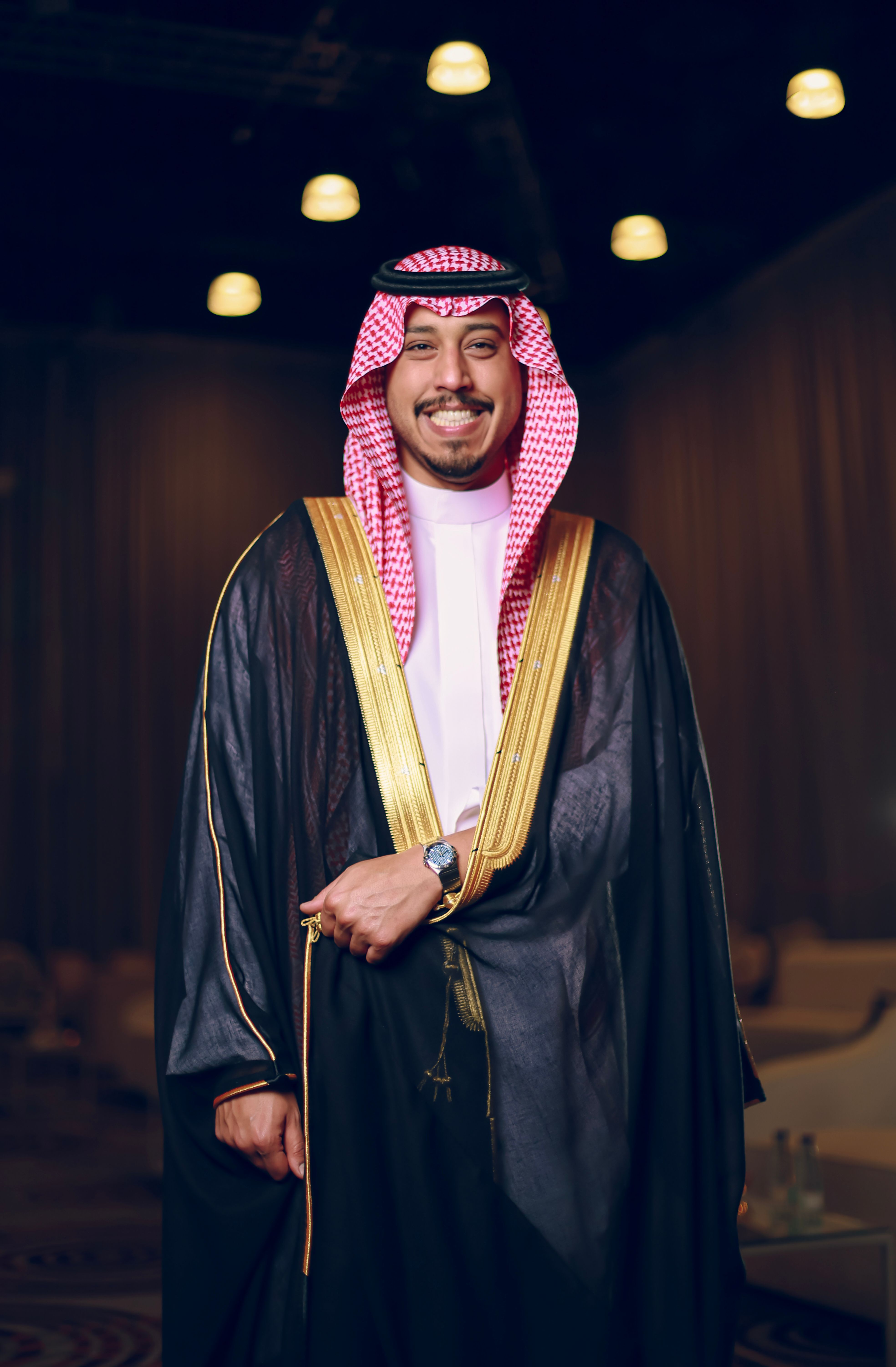 A smiling Middle Eastern man wearing traditional clothing stands indoors under soft lighting.