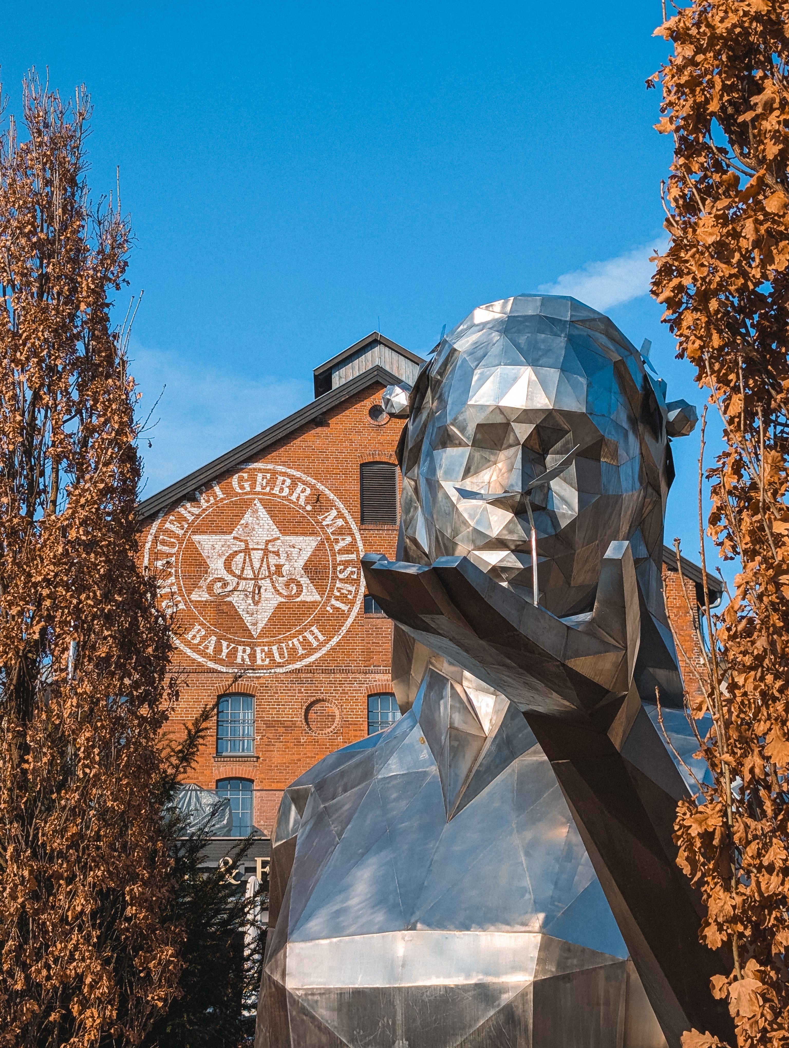 A geometric sculpture stands in front of a historic brewery building in Bayreuth under a clear sky.