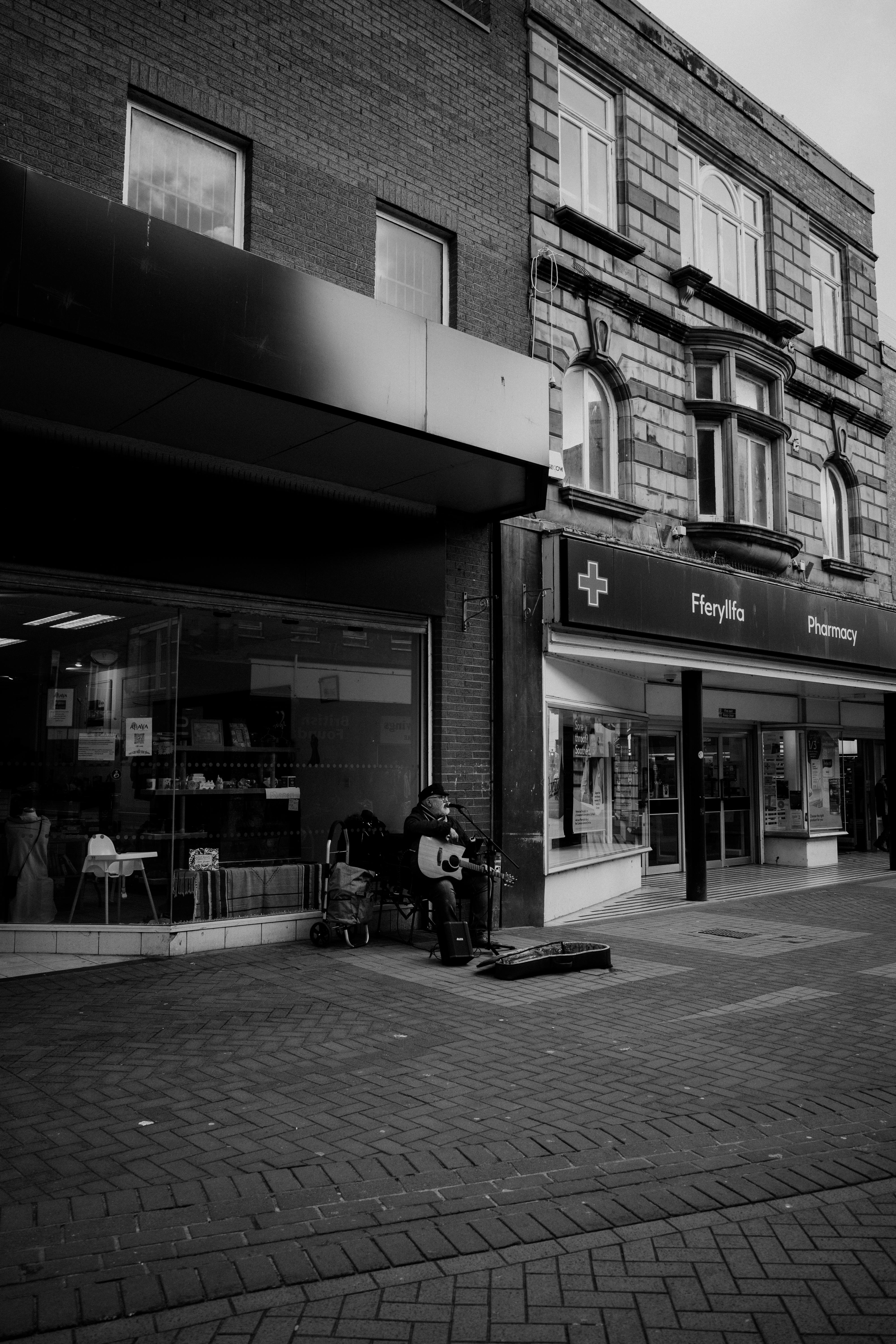 Black and white street scene with musician playing guitar in front of pharmacy.
