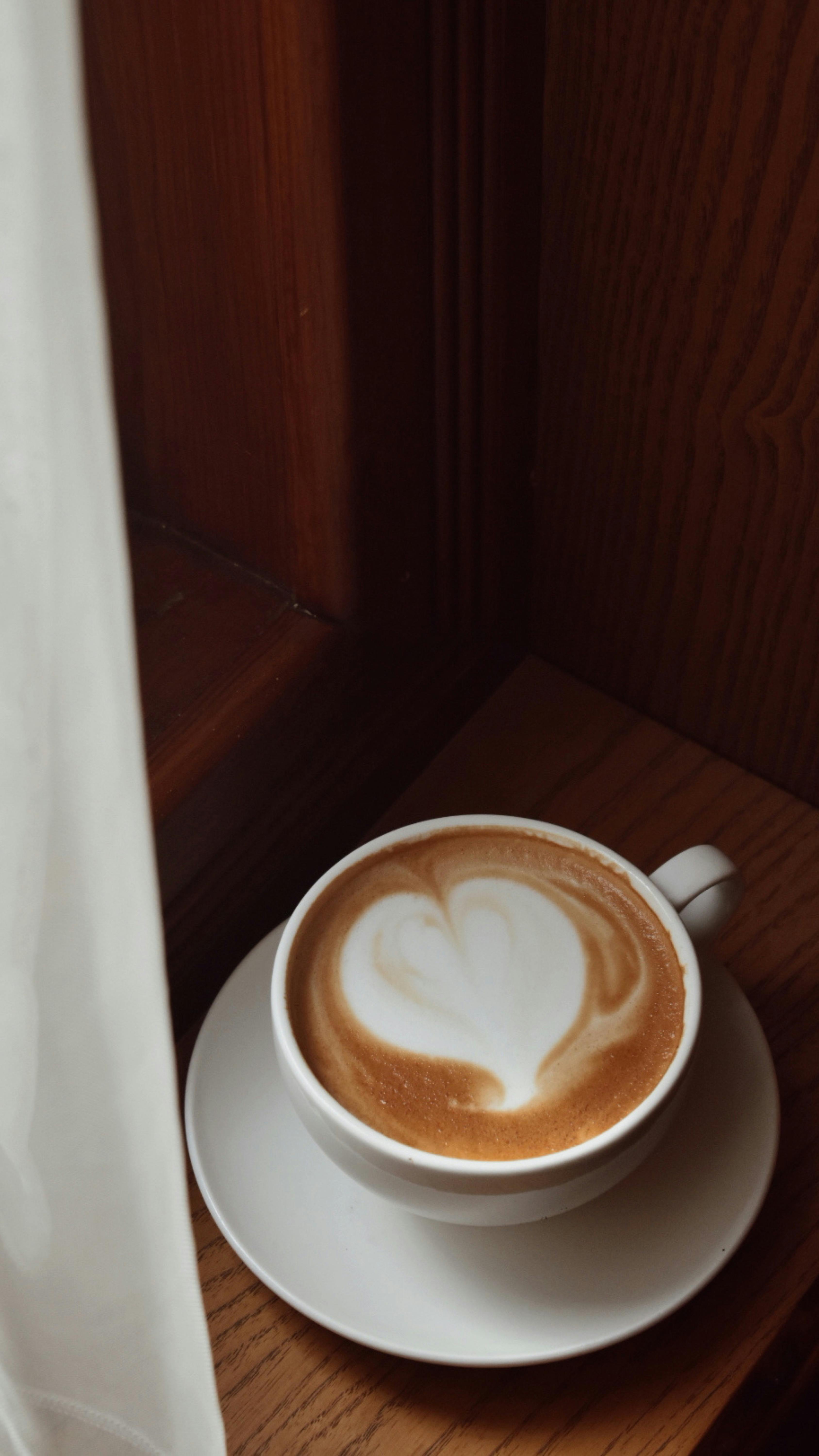 A cappuccino with heart latte art on a wooden table by a window.