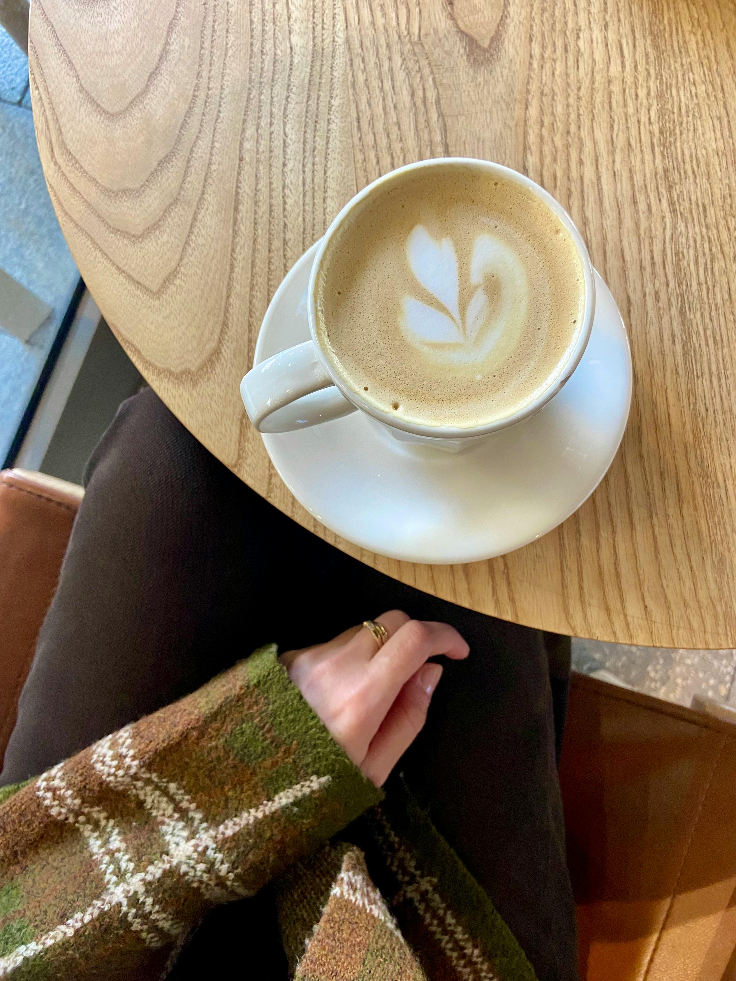 Warm cup of cafe latte with heart latte art on a wooden table beside a person in a knit sweater.
