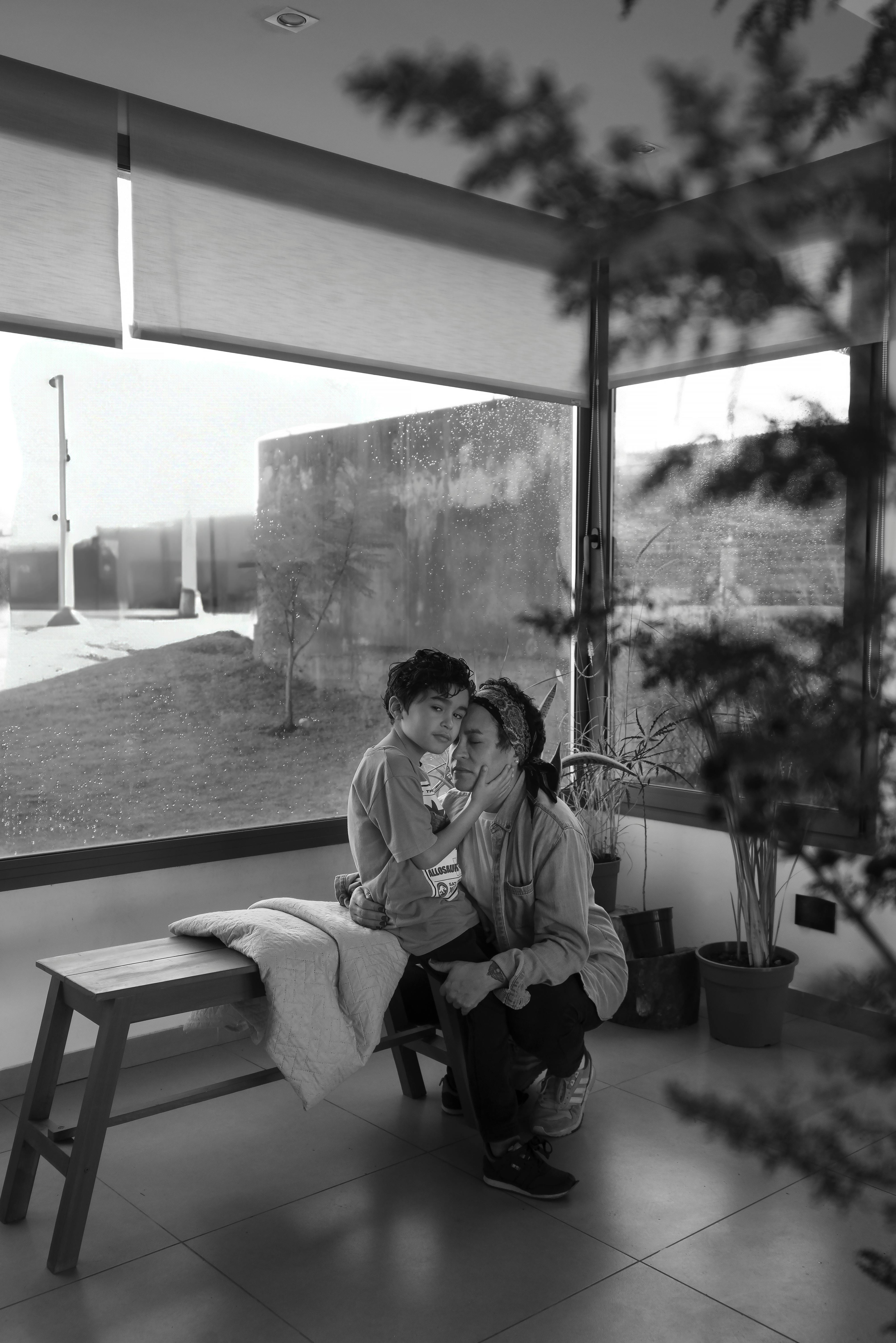 Black and white photo of a mother embracing her child by a window, creating a warm family moment.
