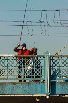 Two men enjoy fishing from a bridge under a clear sky, holding up catches.