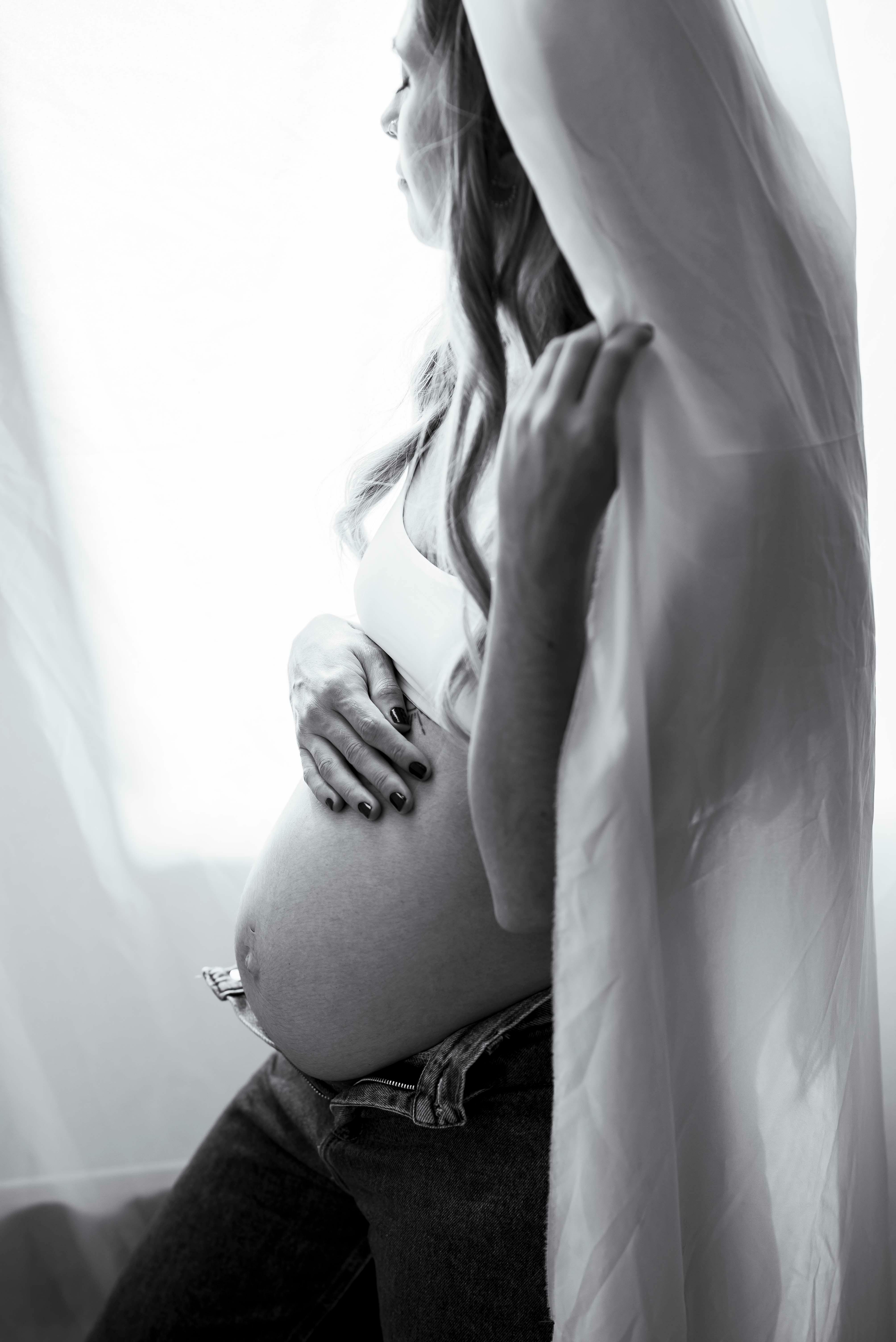 Kostenlos Elegantes Schwarz-Weiß-Porträt einer schwangeren Frau, die ihren Bauch an einem Vorhang festhält. Stock-Foto