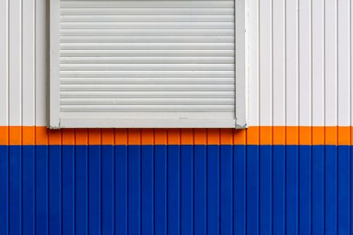 Free A minimalist facade featuring a shuttered window with vibrant blue and orange stripes. Stock Photo