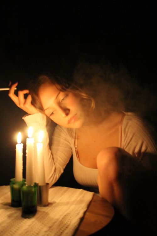 Free A woman smokes pensively by candlelit table, surrounded by darkness. Stock Photo