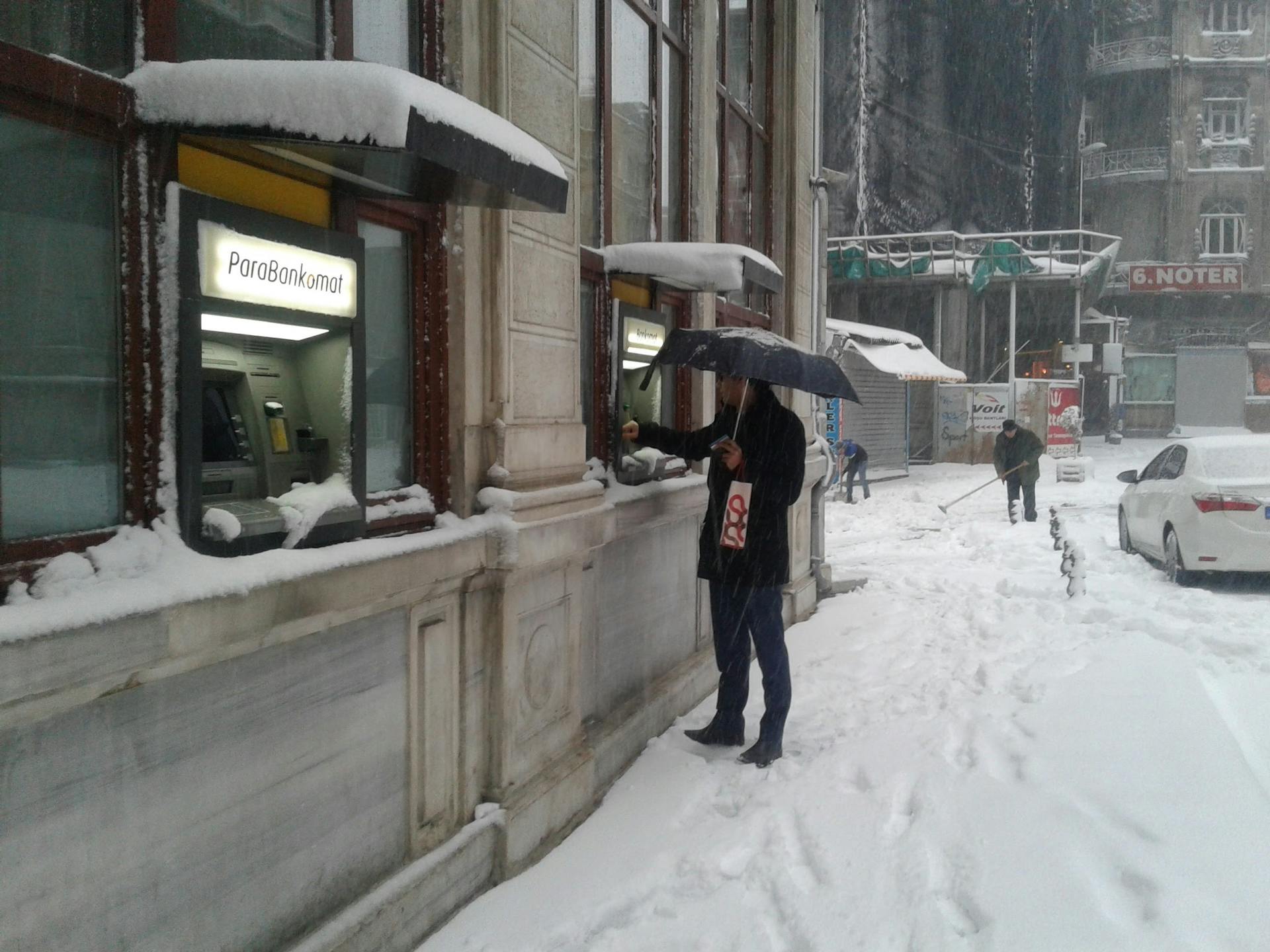 Person using ATM in snowy urban setting, umbrellas out, snowy street.