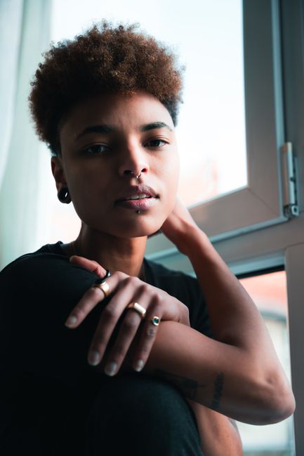 Aesthetic portrait of a young woman with piercings and tattoos by a window in Berlin.