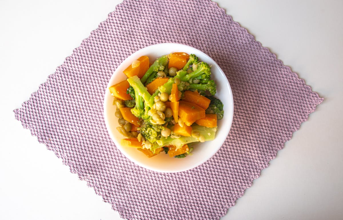 A vibrant mix of cooked vegetables including broccoli and carrots, served in a bowl on a textured mat.