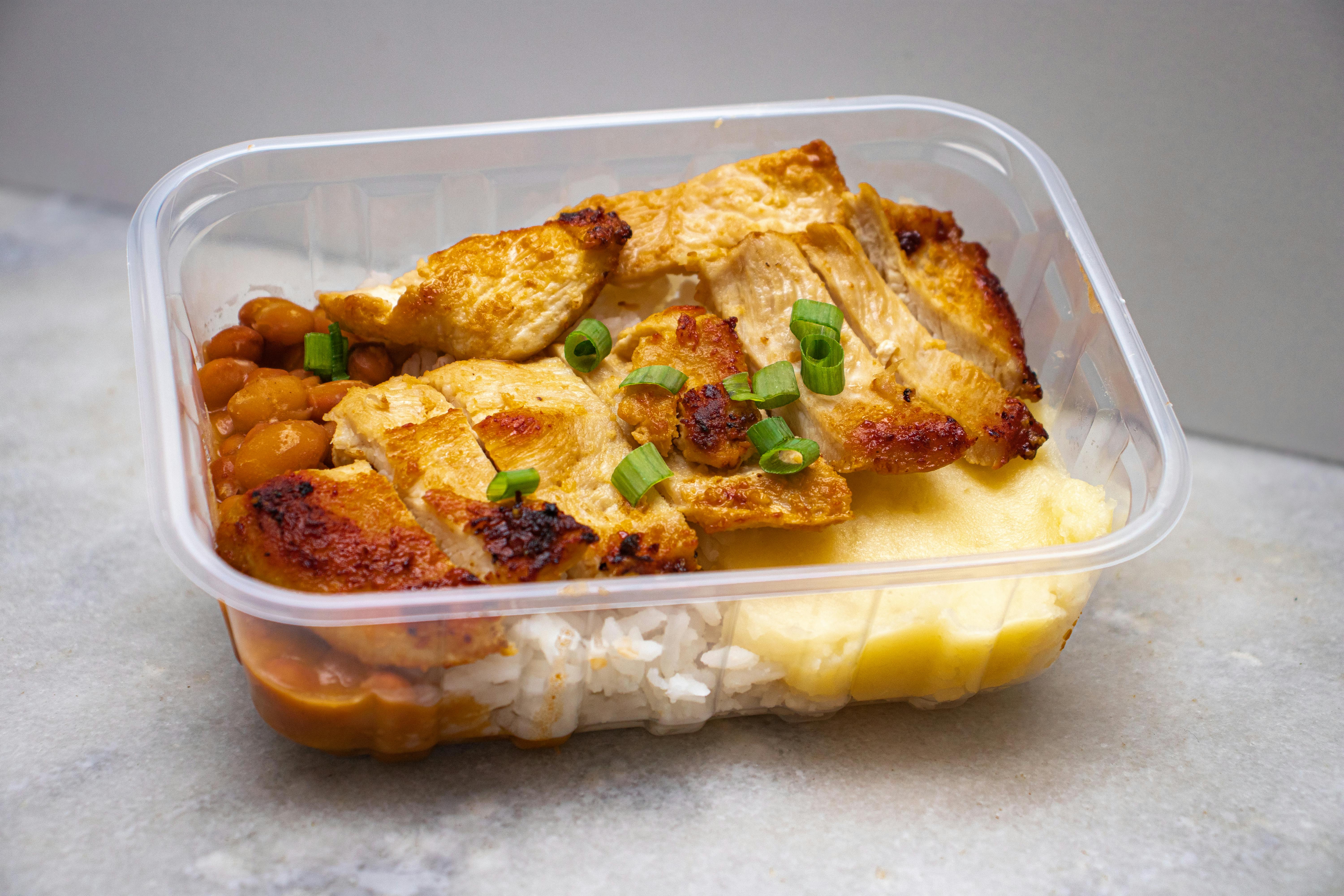 Grilled chicken with rice, beans, and greens in a clear takeout container.