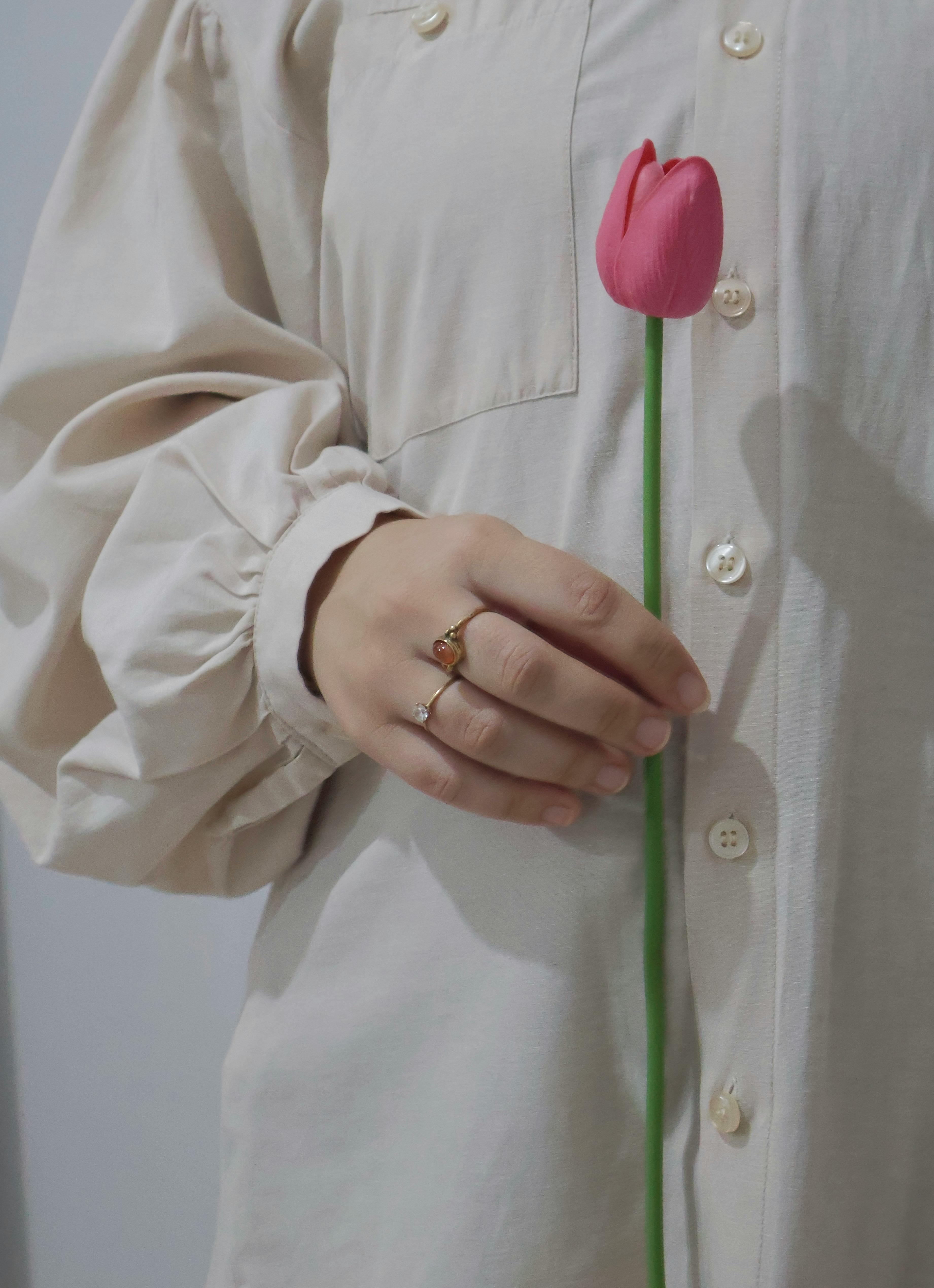 Close-up of a hand holding a pink tulip, wearing a beige shirt and ring. Minimalist style.