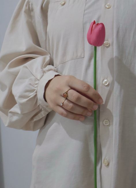 Close-up of a hand holding a pink tulip, wearing a beige shirt and ring. Minimalist style.