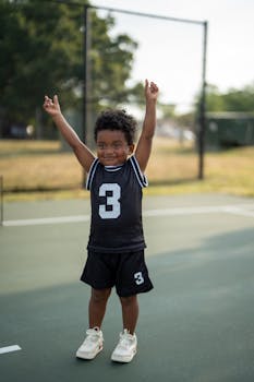 Happy child in a black basketball jersey on an outdoor court, raising arms in excitement.