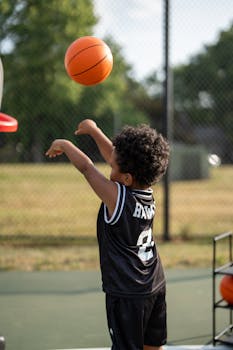 A young boy in a black jersey shoots a basketball on an outdoor court, enjoying a sunny day.