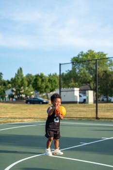 A cute child in a black jersey holding a basketball on an outdoor court.