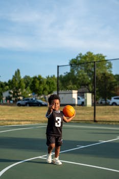 Happy toddler in black jersey playing basketball outdoors on a sunny day.