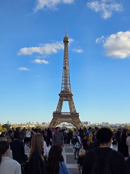 Free A busy day at the Eiffel Tower with crowds gathered under a vibrant blue sky. Stock Photo