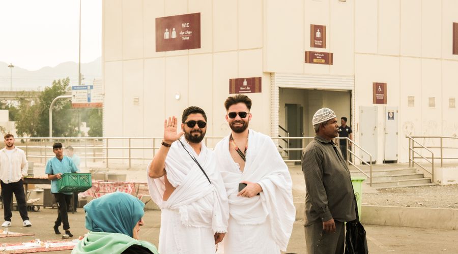 Two male pilgrims wearing white ihram sheets and a small crossbody bag outside a building in Makkah