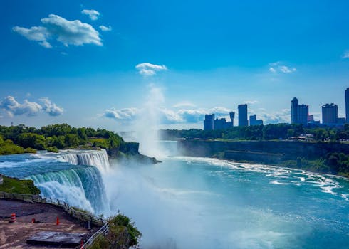 Breathtaking image of Niagara Falls with mist and cityscape under a clear blue sky. by Salah Alawadhi