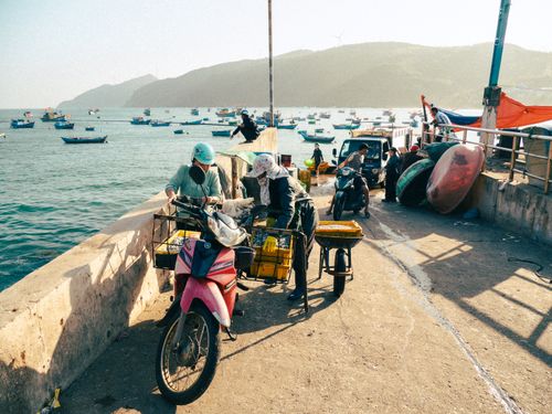 Free Fishermen at Quy Nhơn port loading goods onto motorcycles with boats in the background. Stock Photo
