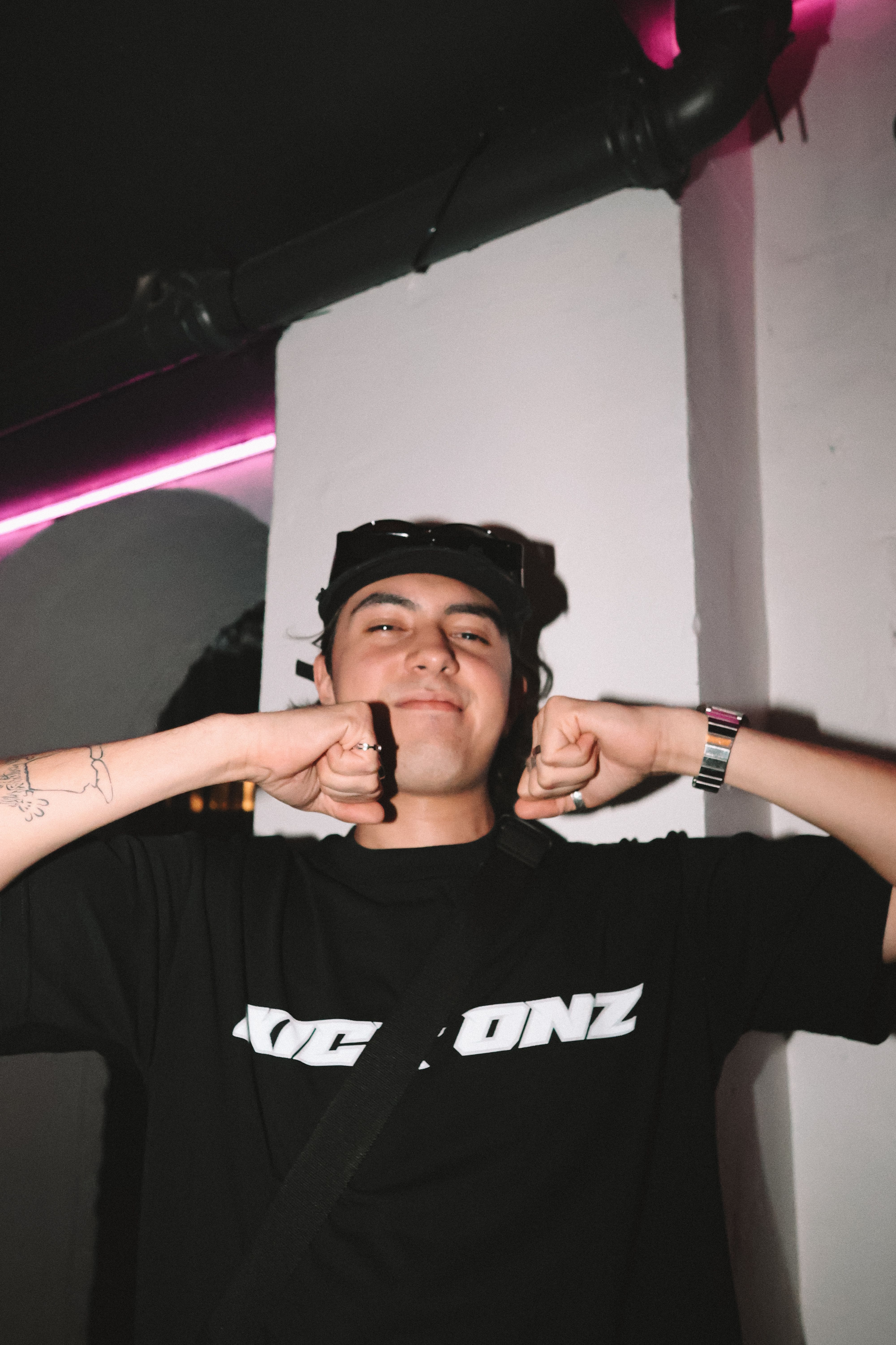 A young adult poses happily in a dimly lit club environment with vibrant neon lights.