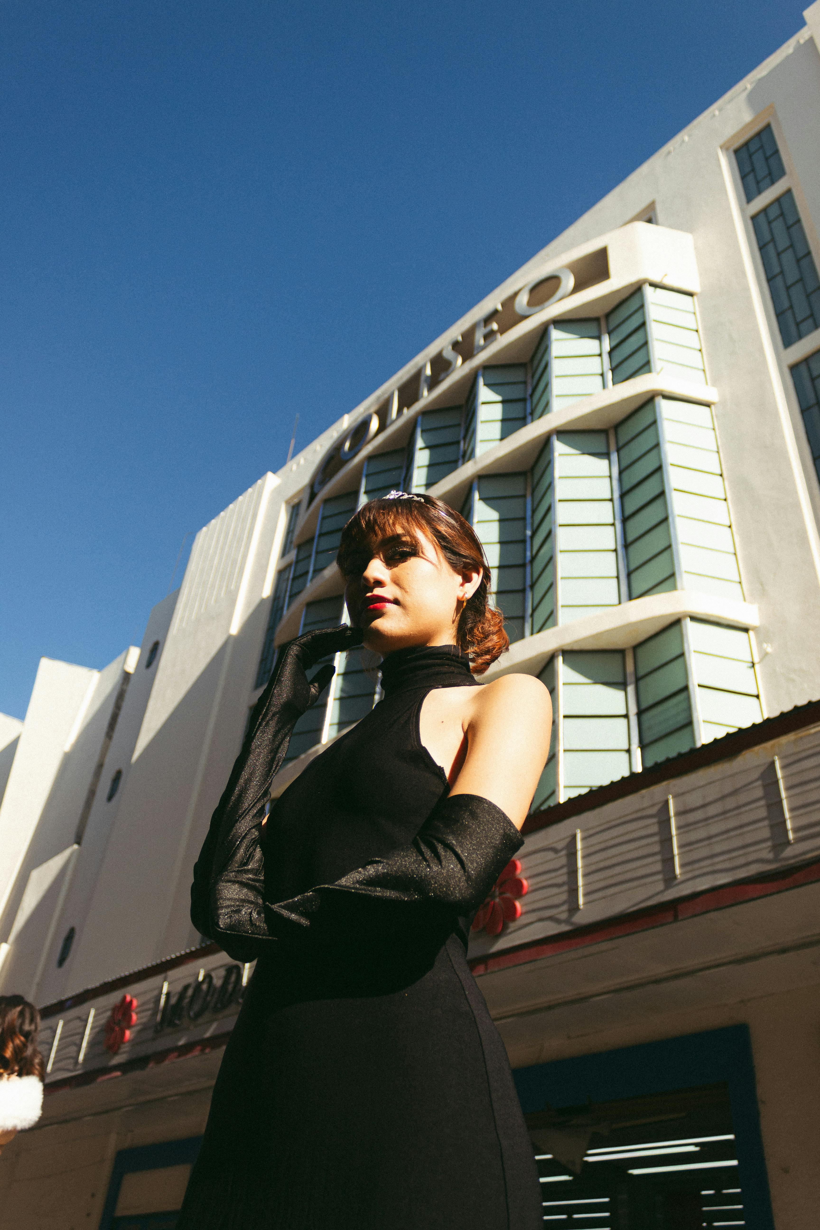 Elegant woman in black dress and gloves poses in front of an Art Deco building on a sunny day.
