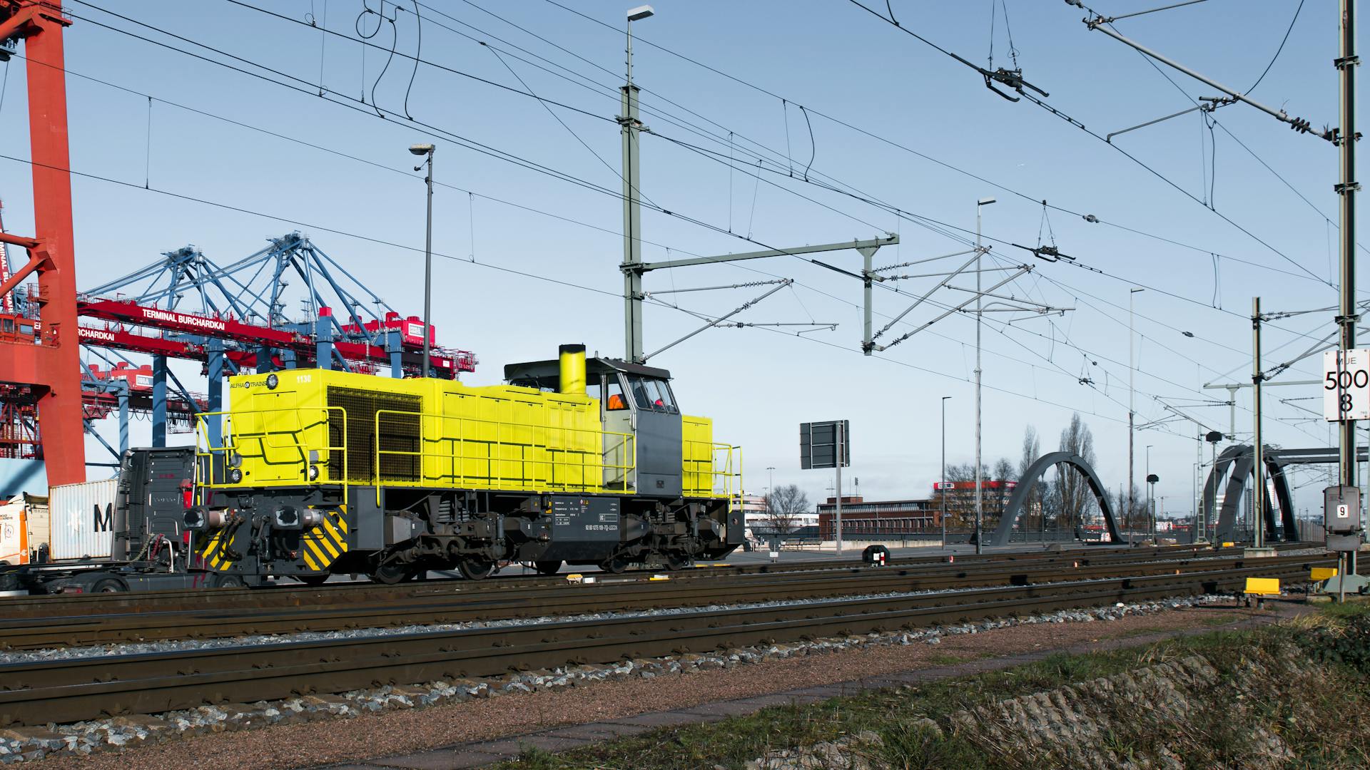 A striking yellow locomotive at Hamburg port amidst shipping containers and rail infrastructure.