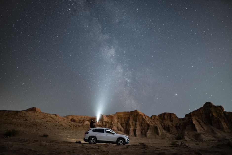 A person stands on an SUV under a vast starry night sky in the desert, amid rock formations.