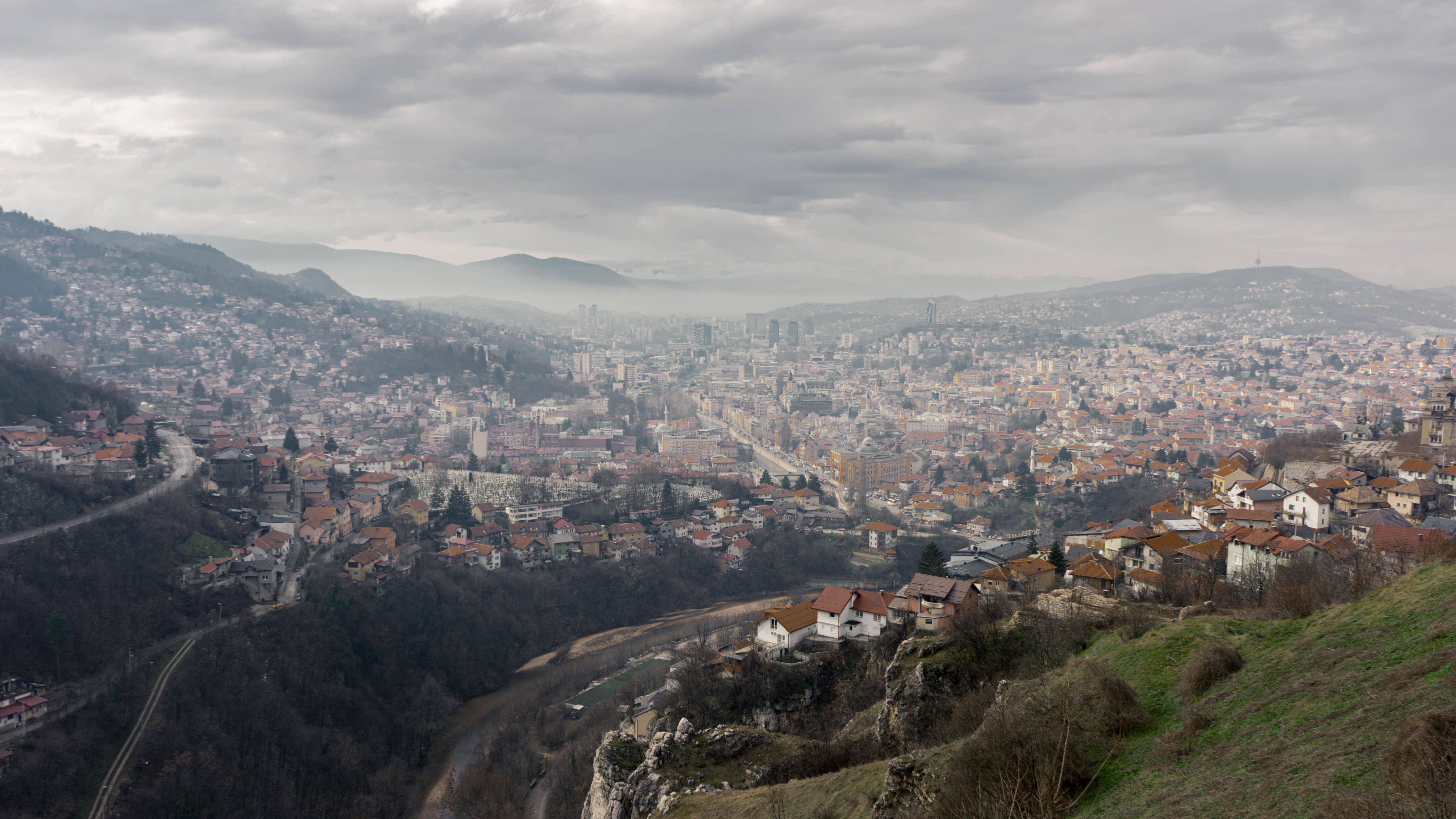 A panoramic view of Sarajevo under overcast skies. Hills and city life converge.
