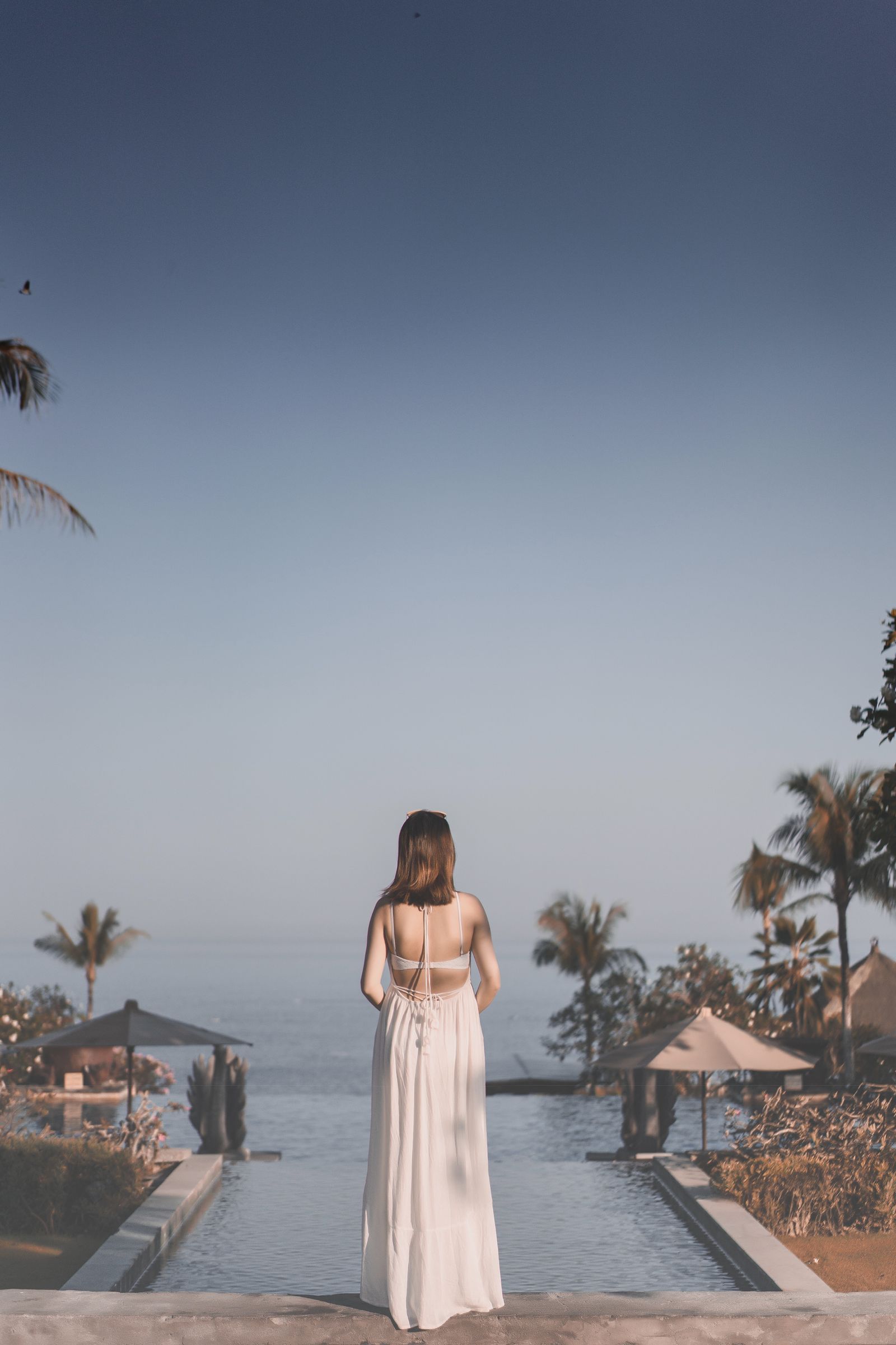 Woman wearing an elegant white dress overlooking a tropical infinity pool