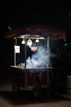 Street vendor roasting chestnuts at night, surrounded by smoke and illuminated by lamps.