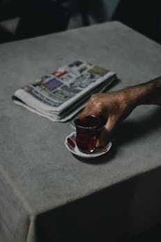 A hand holding a traditional Turkish tea on a table with a folded newspaper.