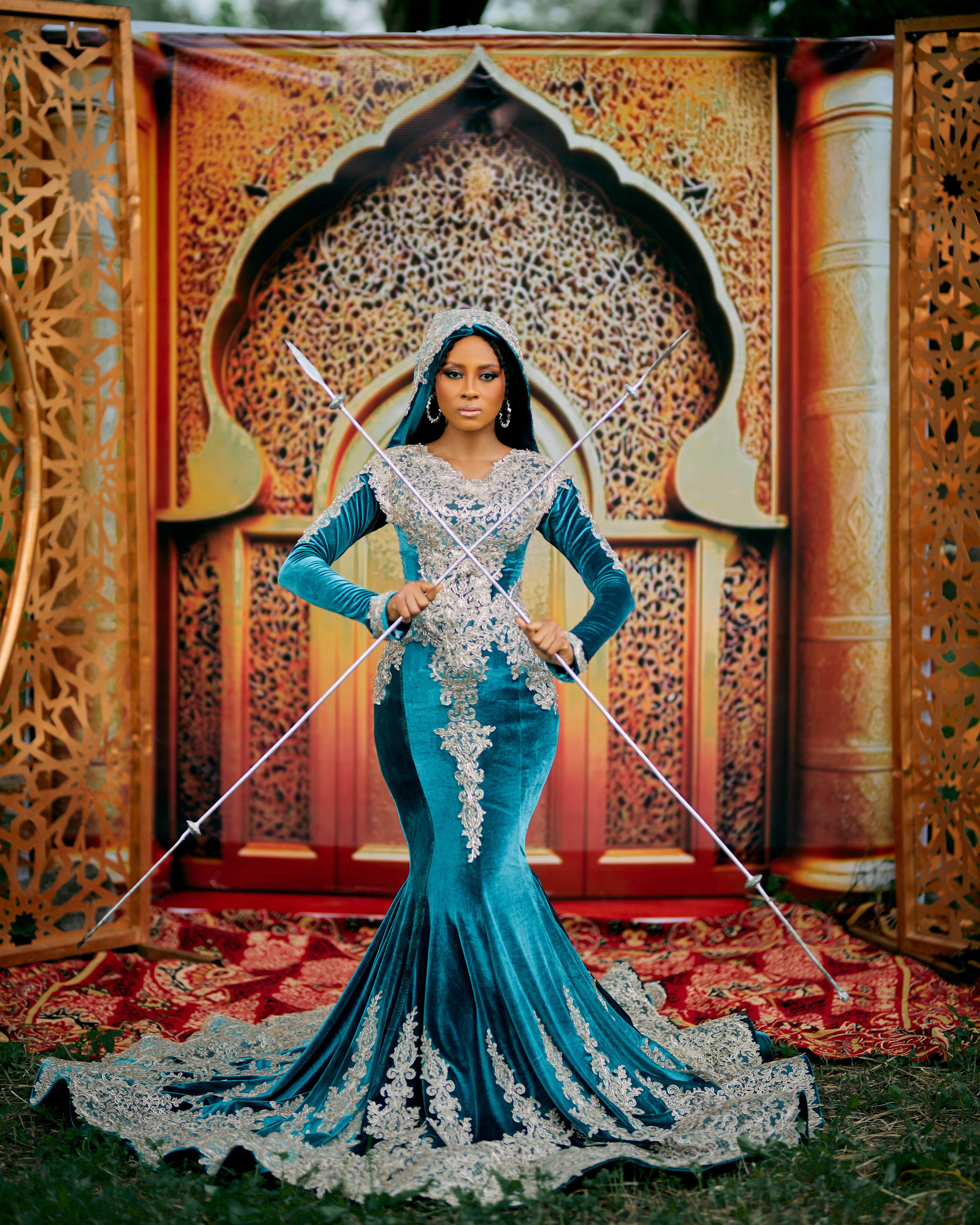 A woman in a blue and silver gown stands before an intricately designed Arabic backdrop.