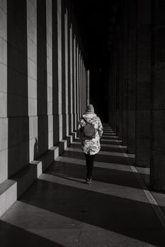 A woman walks through a sunlit corridor in black and white, highlighting shadows and architectural columns.