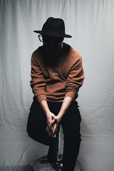 Moody portrait of a man in a hat and sweater sitting against a soft white backdrop.