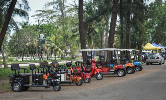 Row of colorful electric scooters and golf carts parked under trees in a park.