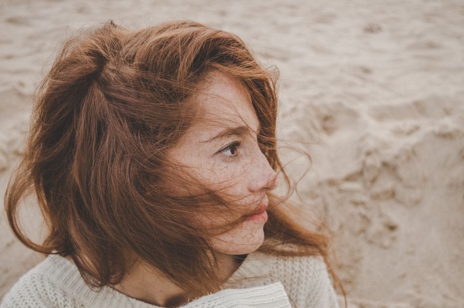 Red-haired woman with freckles on a sandy beach, gazing thoughtfully. Natural beauty and serene atmosphere.