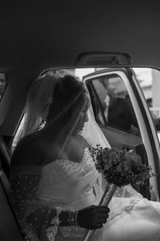Black and white photo of a bride in a car with bouquet, radiating elegance and anticipation.