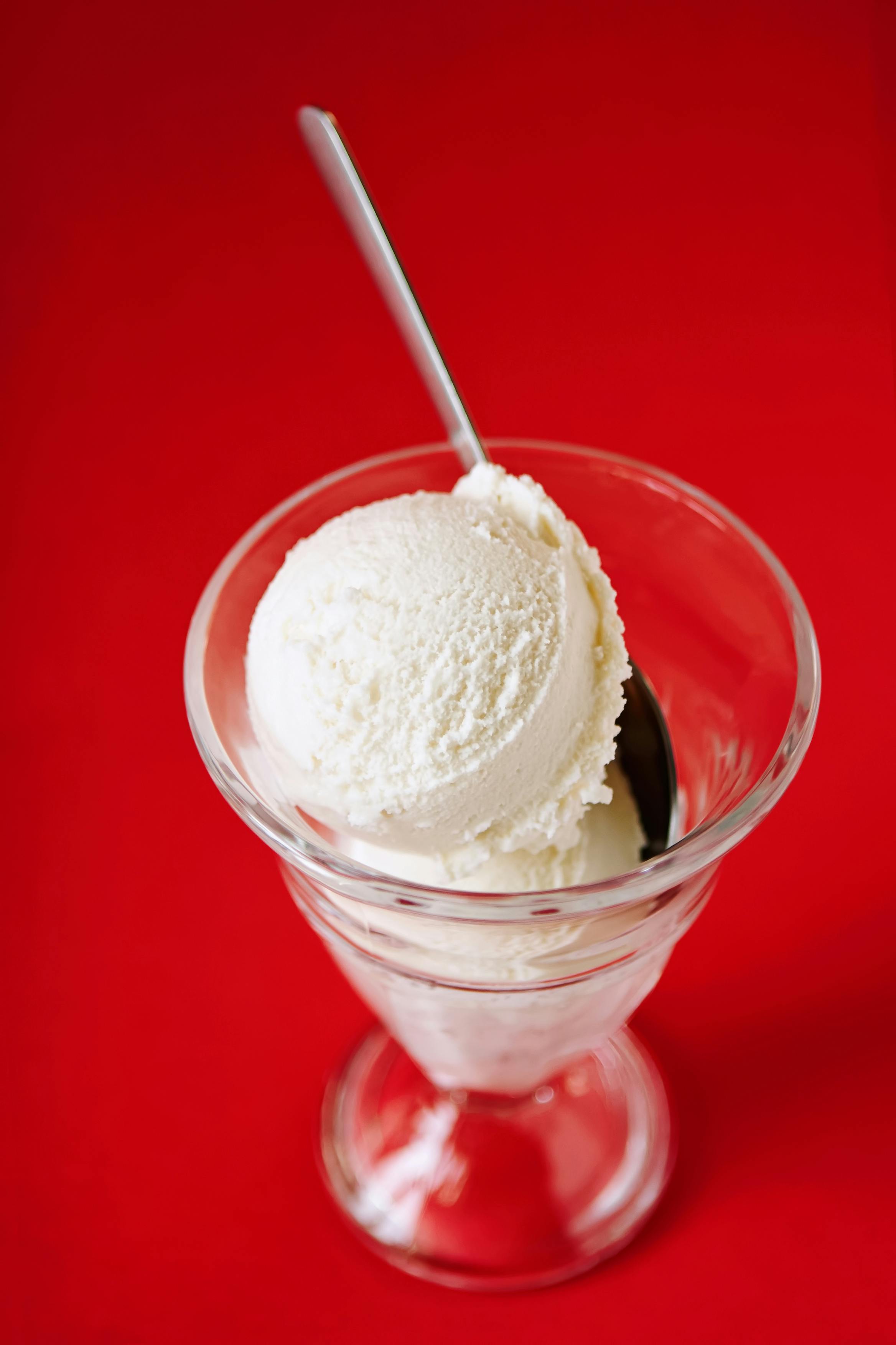 Delicious vanilla ice cream scoop in a glass dish with spoon, set against a vibrant red backdrop.