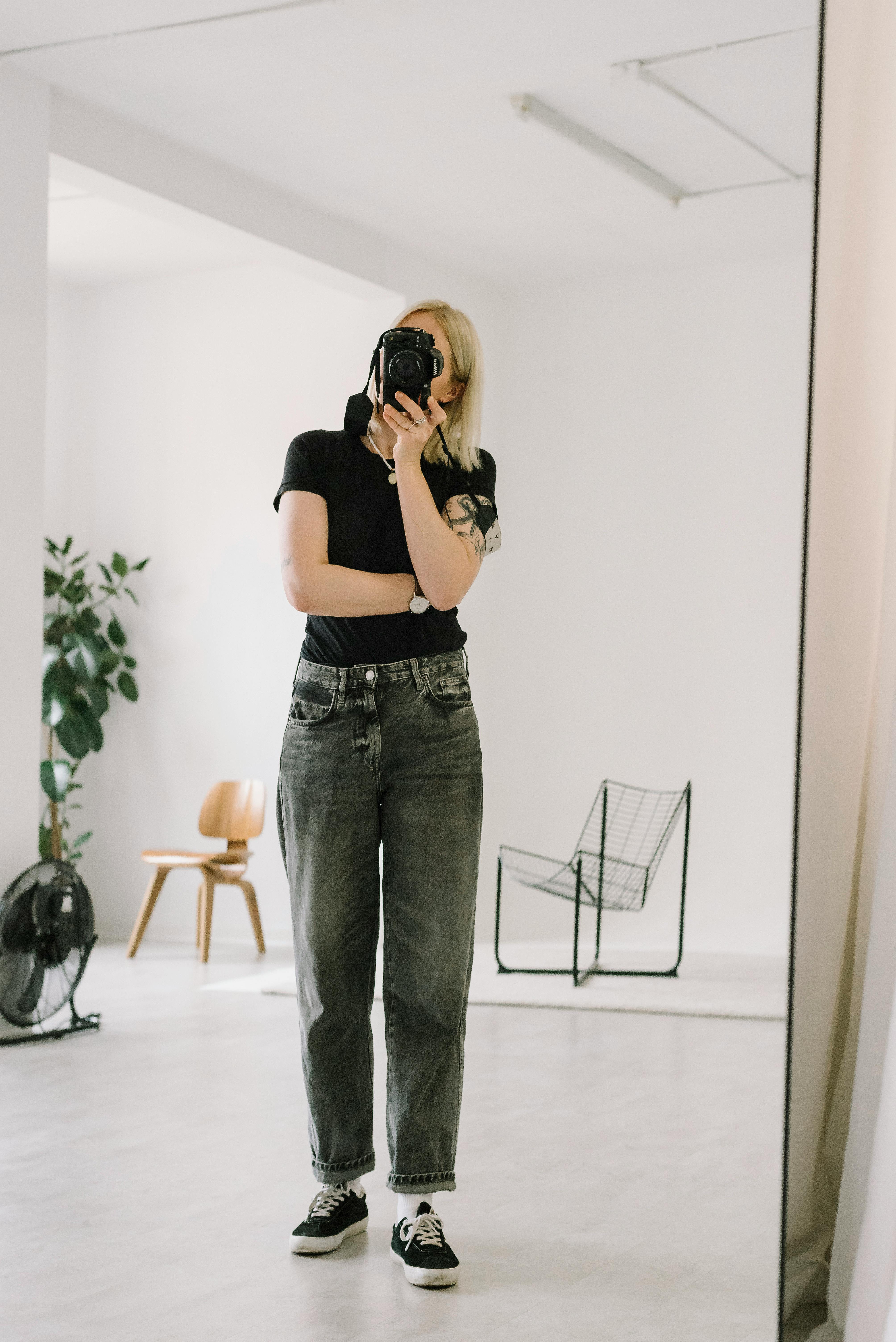 A person takes a mirror selfie in a bright, minimalist studio with natural elements.