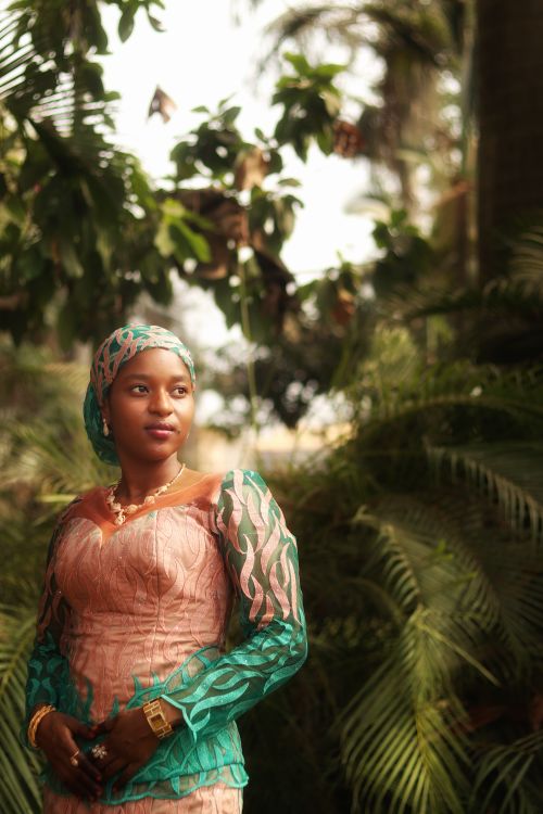 Free A woman in vibrant Nigerian attire stands gracefully against lush greenery, showcasing cultural elegance. Stock Photo