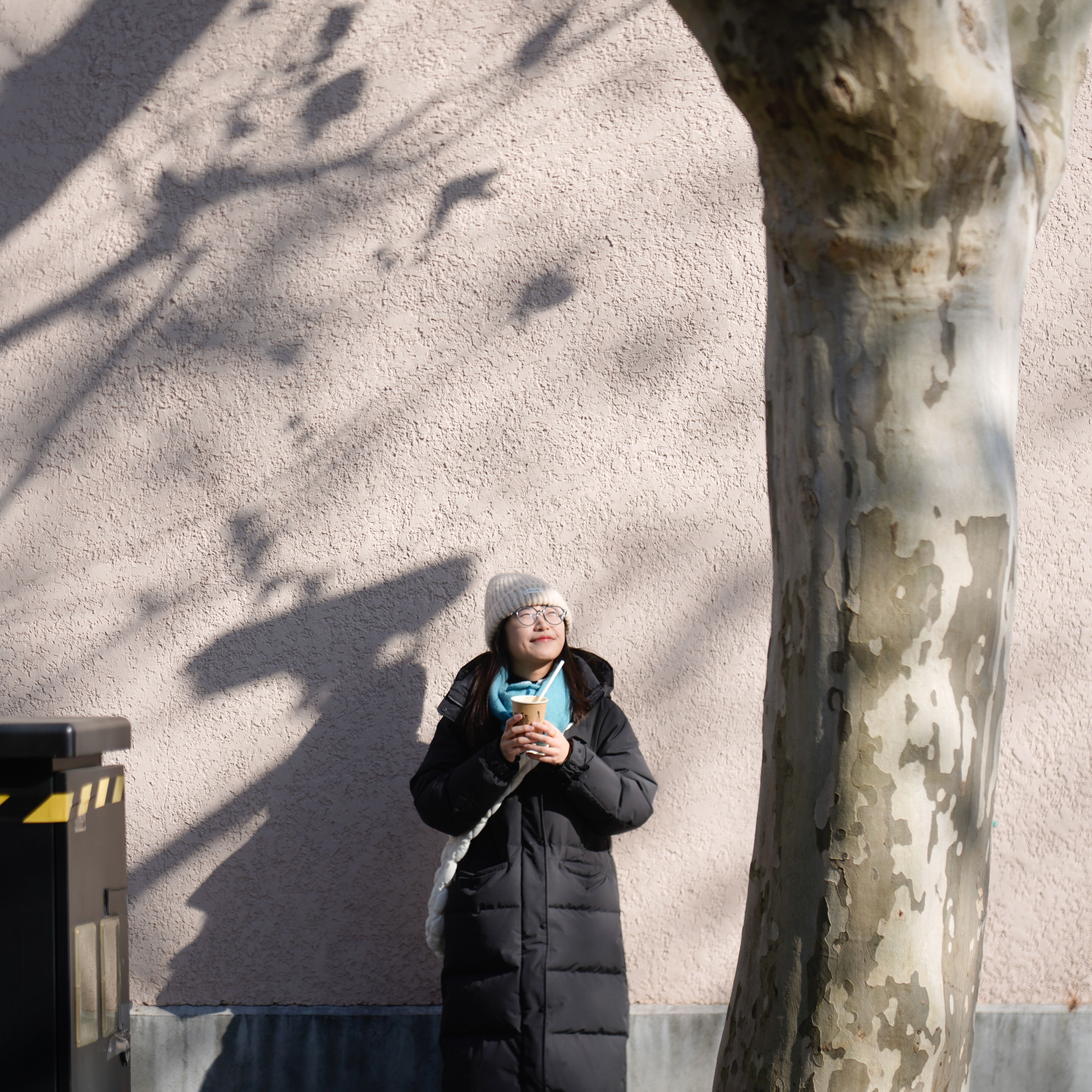 A woman in a winter coat enjoys a warm drink against a sunlit wall.