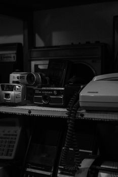 A moody black and white image of vintage cameras and electronics on a shelf.