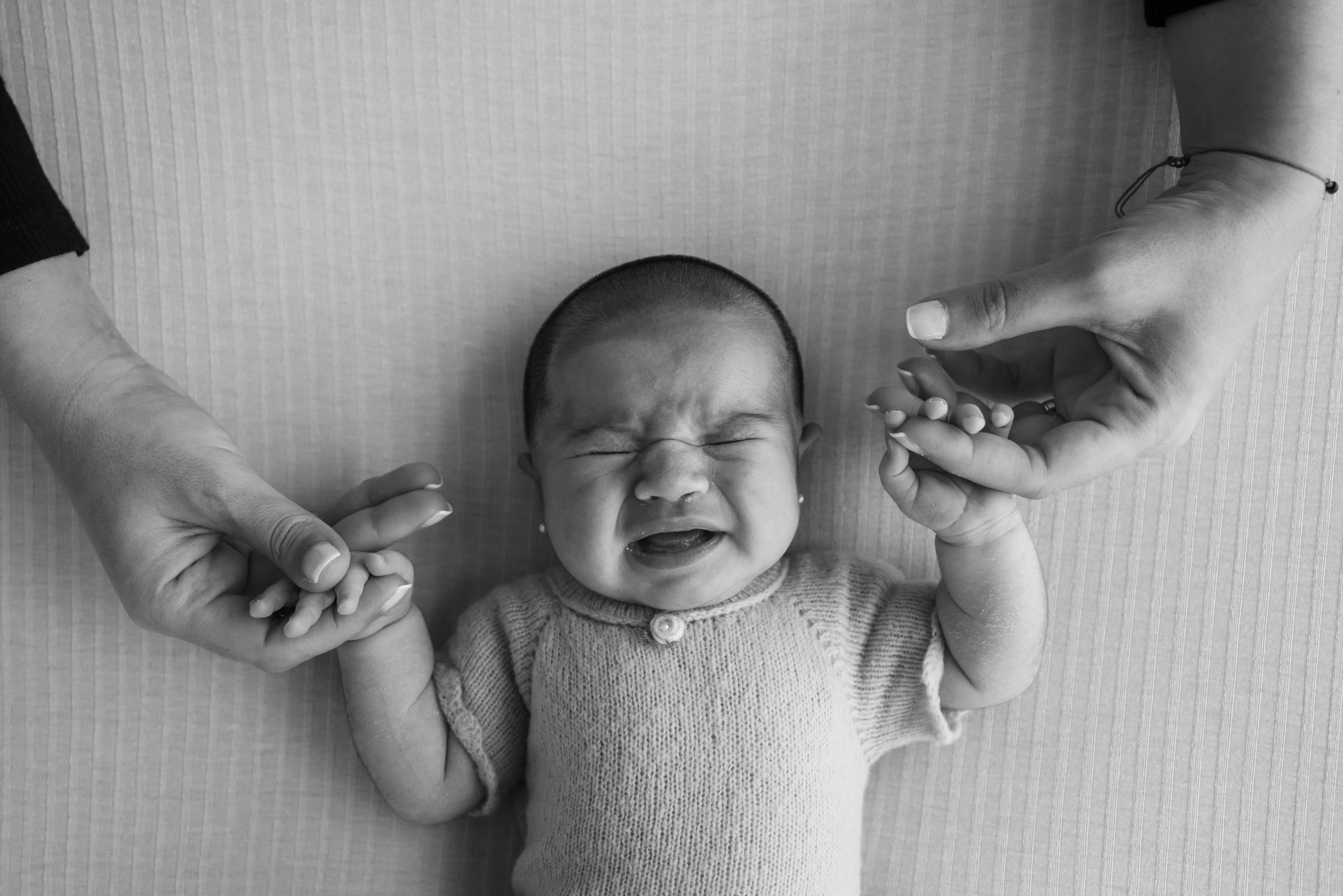 Black and white image of a crying baby being comforted by two adult hands holding theirs.