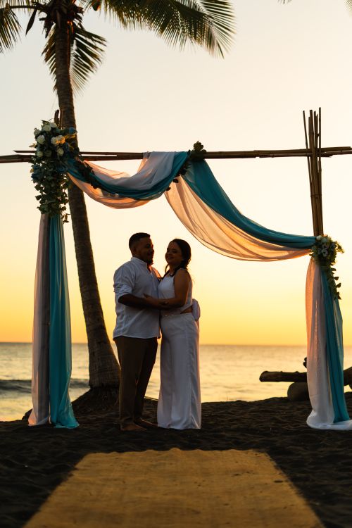 Free A romantic beach wedding ceremony with a couple embracing under a decorated arch at sunset. Stock Photo