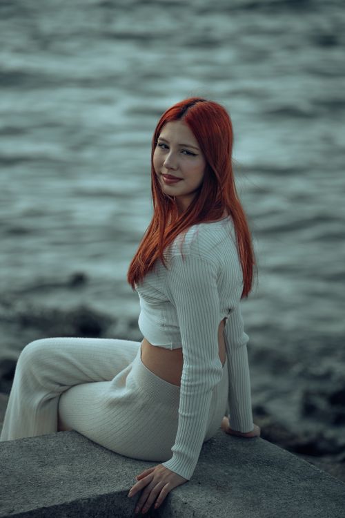 Free Portrait of a young woman with red hair sitting by the waterside edge. Stock Photo