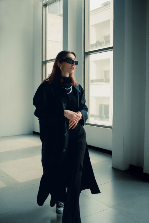 Free Fashionable woman in a black coat and sunglasses walking indoors near large windows. Stock Photo
