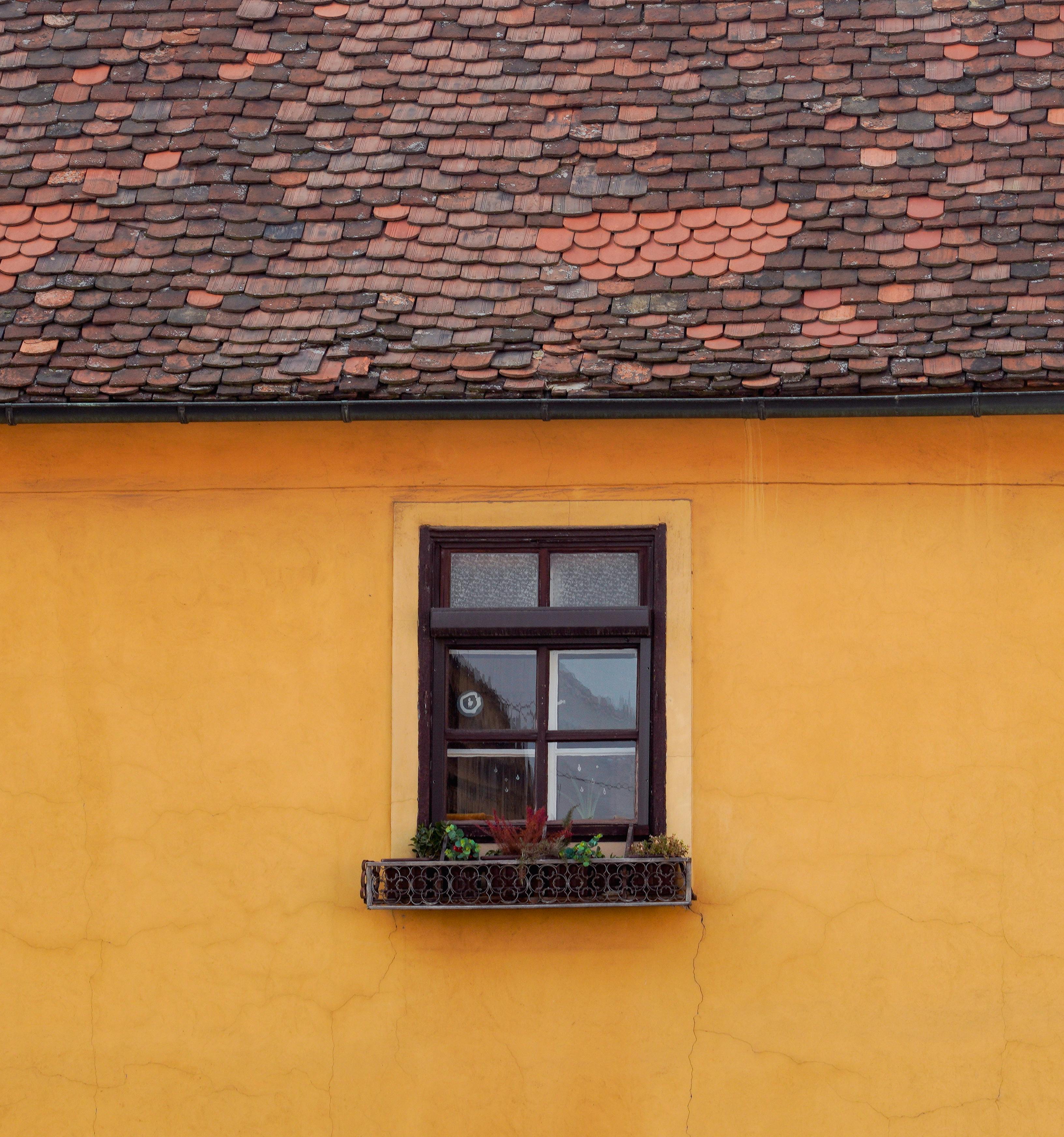 Rustic window on a yellow wall with vibrant roof tiles and a quaint flower box.