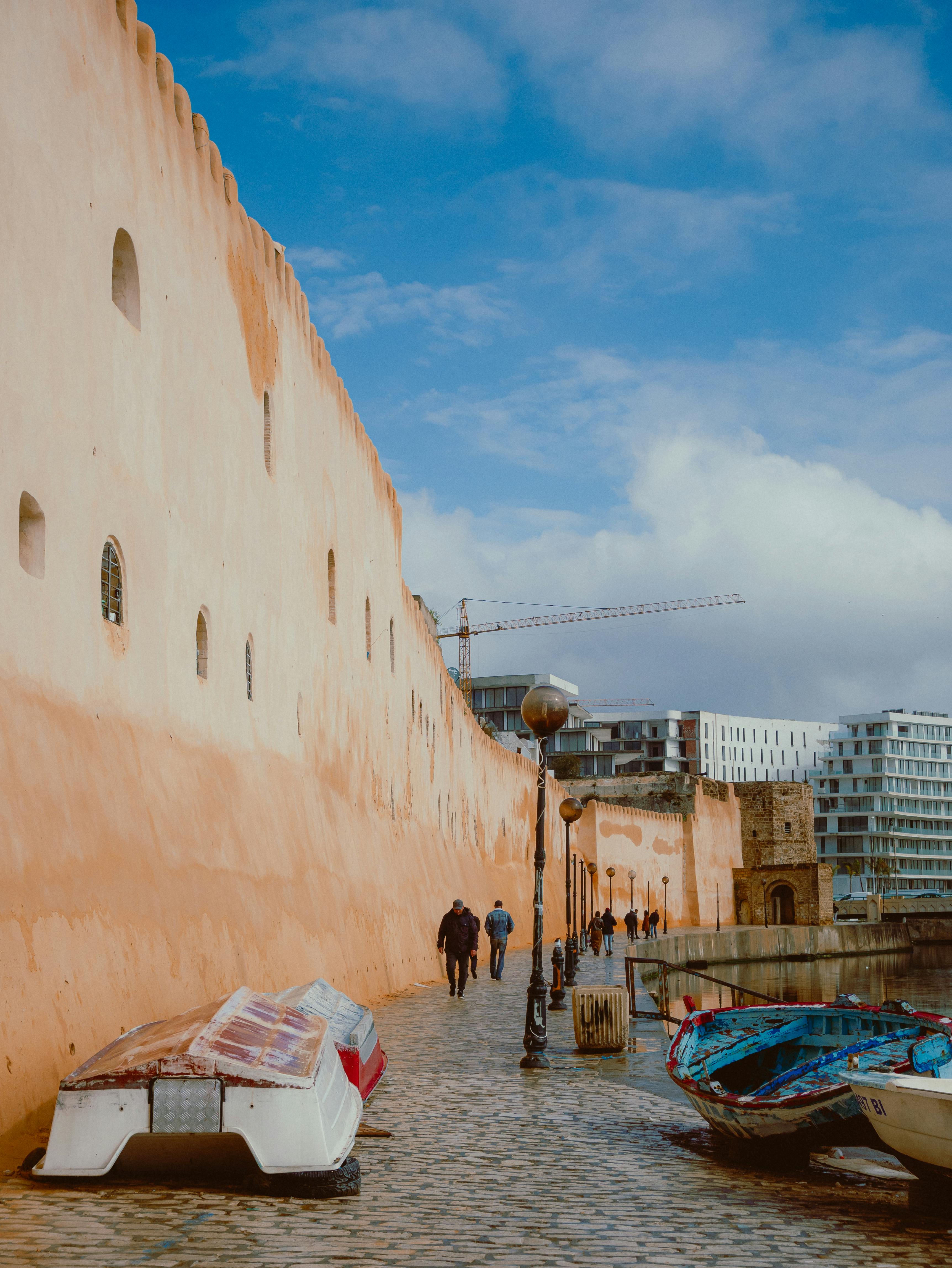 People walking along a historic coastal path with boats and cityscape in the background.